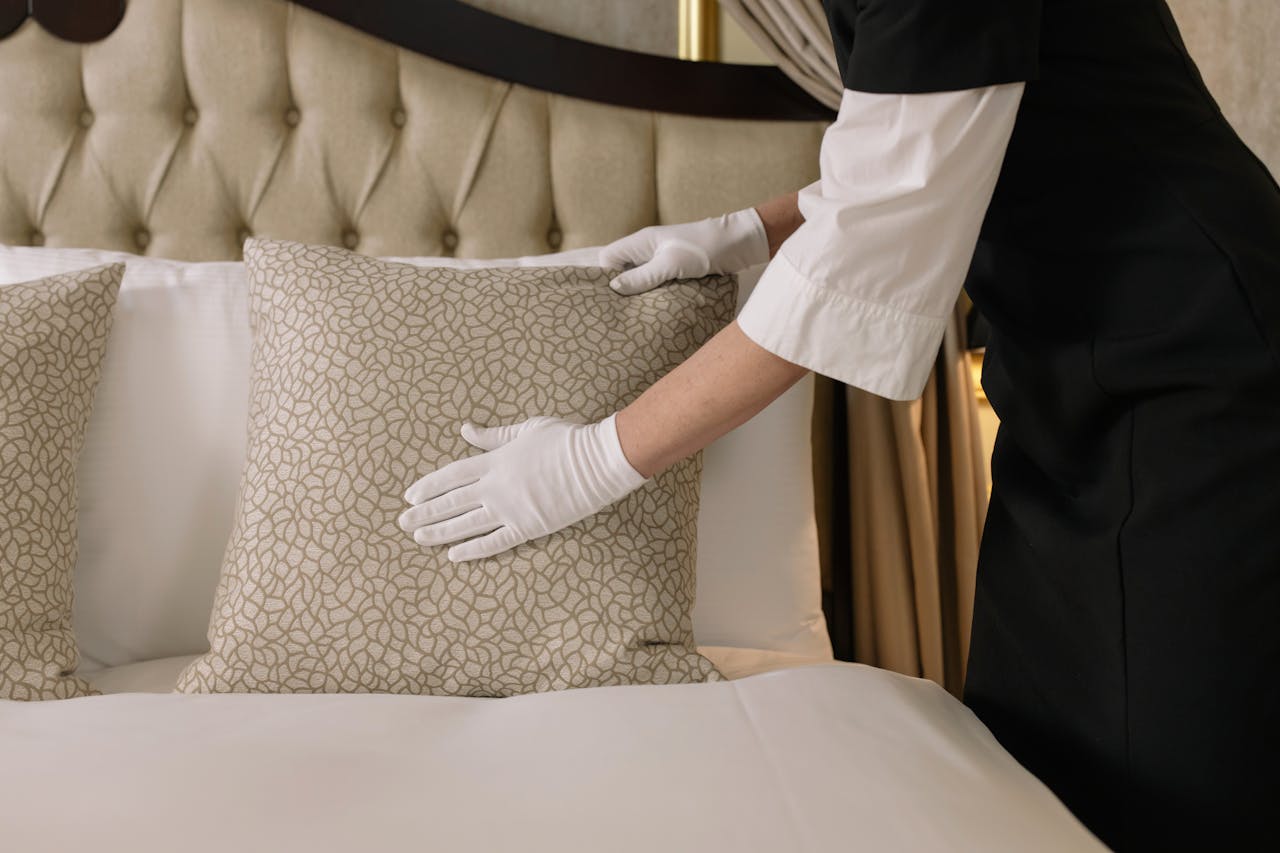 services-01 A housekeeper wearing gloves arranges pillows on a luxurious bed in a hotel room.